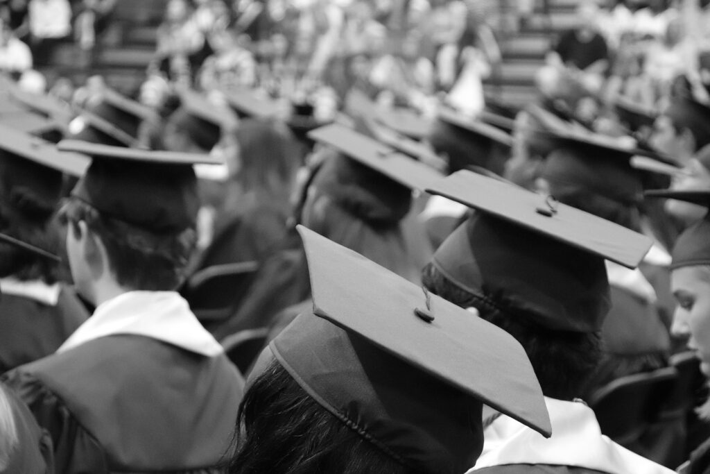 Black and white photo of a crowd of graduates in caps and gowns, viewed from as though standing among them, looking at the backs of their heads toward a stage and audience in the bleacher seats.
